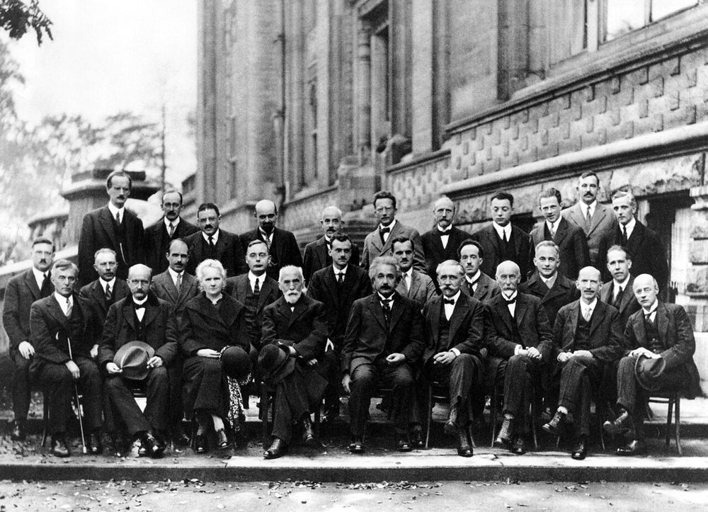 A black-and-white group photograph of 29 scientists in formal dress, standing and seated in three rows in front of a building.